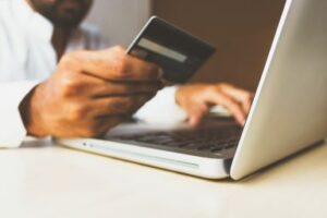 A picture of a man holding a blue credit card while working on a silver laptop. Buying from a Charity Auction? Custom Development Solutions. 