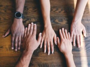 A group of people put their hands on a brown table. Their skin color is progressively darker form right to left. Philanthropy has no color. Custom Development Solutions.