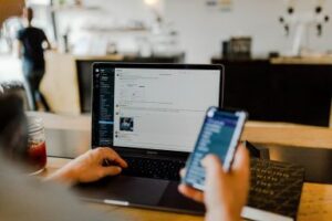 A picture of a man using both his phone and his computer togeheter in an office. One must design a Communications plan. CDS Fundraising 