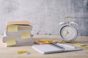 Books, notepads, a notebook, and a clock on a desk representing how to manage your time more efficiently.