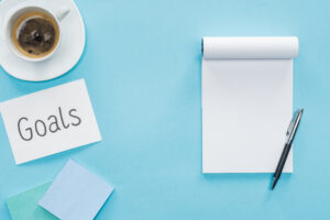 Top view of a cup of coffee, a blank notebook, and a card that says "goals." It is important to set realistic goals in your fundraising!