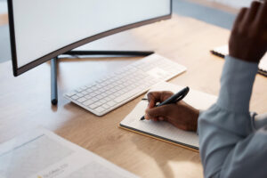 Business worker in an office writing a compelling case statement in a notebook in front of a monitor. 