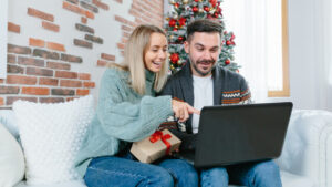 This photo is of a young couple sitting on a sofa in front of a Christmas tree as they do their holiday shopping together on a laptop.