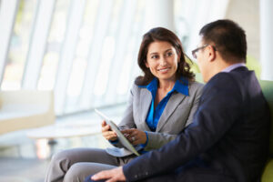 A smiling woman in business attire talking to a man and pointing to something on a tablet